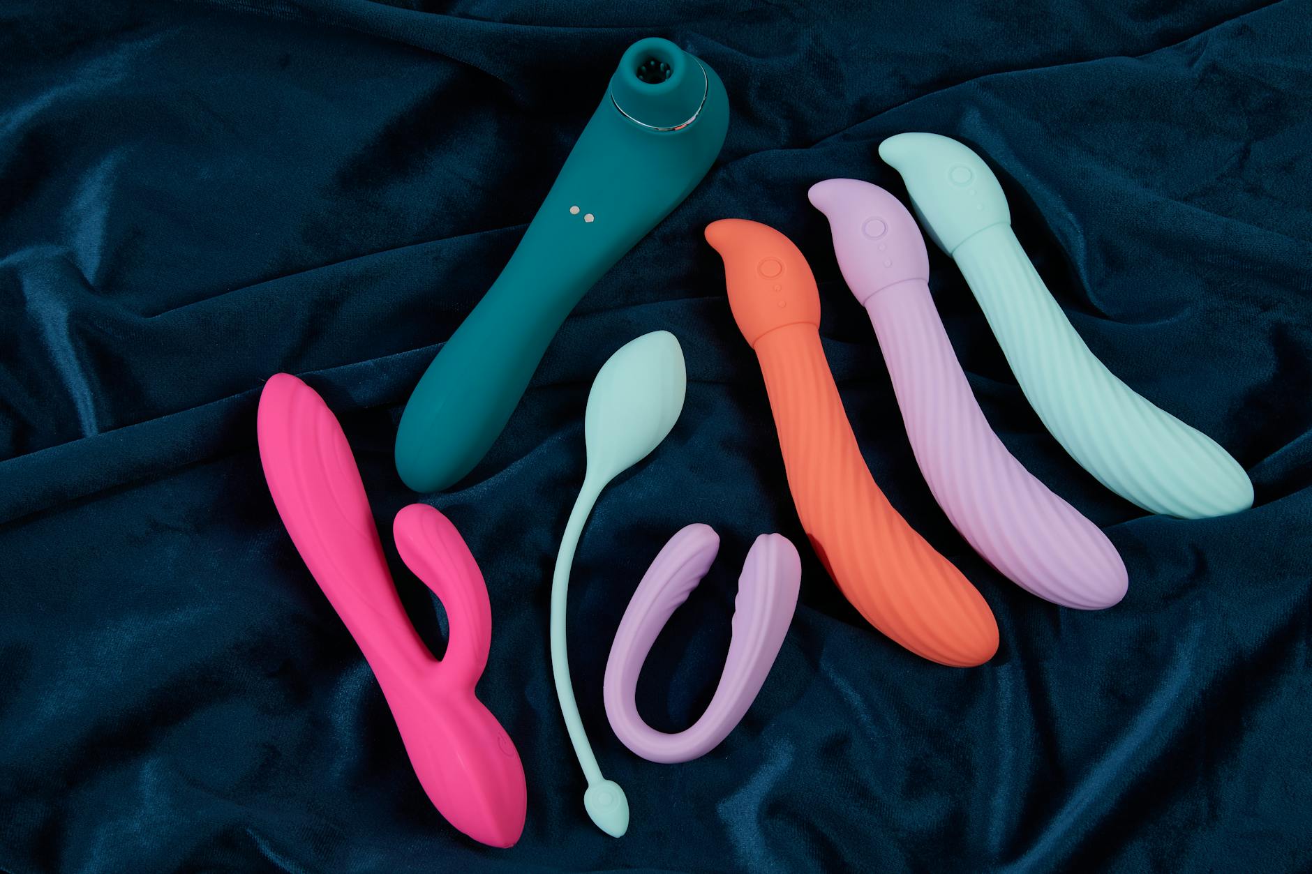 Display of colorful vibrators and clitoral stimulators arranged on dark blue fabric, showing various shapes and designs