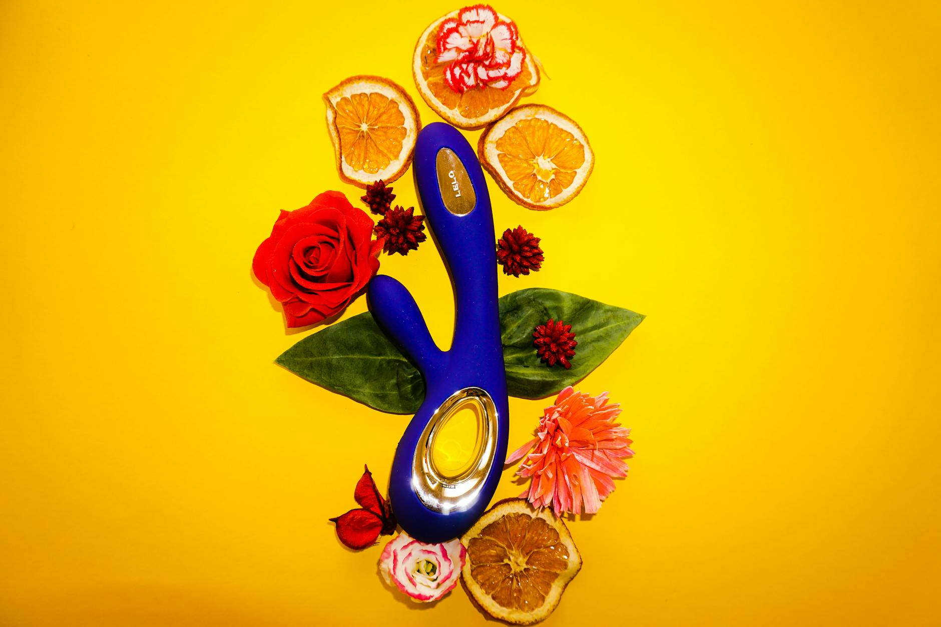 A blue clitoral vibrator surrounded by flowers and citrus on a bright yellow background
